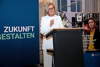 Die niederösterreichische Landeshauptfrau Johanna Mikl-Leitner spricht am Rednerpult bei der Eröffnung des „Tags der NÖ Industrie“ in Laxenburg; im Hintergrund ein Plakat der IV-NÖ-Kampagne „Damit AUT nicht OUT wird“.